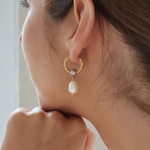 May include: A gold hoop earring with a pearl dangle. The earring is worn by a person with long brown hair.
