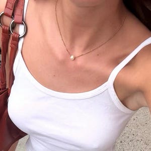 May include: A white sleeveless tank top, a white skirt with a subtle dotted texture, a gold chain necklace with a single pearl pendant, a set of gold bangles, a dark red leather shoulder bag with silver rings, and black ballet flat shoes.