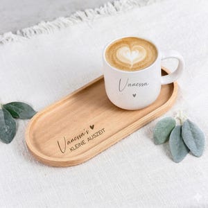 Könnte beinhalten: Eine weiße Keramiktasse mit gesprenkelter Oberfläche und herzförmiger Latte-Art, bedruckt mit dem Namen Vanessa. Die Tasse steht auf einem ovalen Holztablett, das mit dem Text Vanessa's KLEINE AUSZEIT und einem kleinen Herz graviert ist.