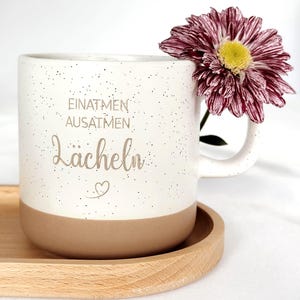 Könnte beinhalten: Eine weiße Keramiktasse mit gesprenkeltem Design und beigem Boden, mit den deutschen Worten "EINATMEN, AUSATMEN, Lächeln" und einem Herzsymbol. Eine weinrote und gelbe Blume ruht auf dem Henkel der Tasse. Die Tasse steht auf einem Holztablett.