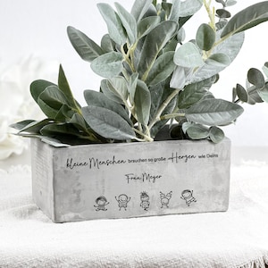 May include: A rectangular concrete planter box with green and gray foliage. The planter has the text "Kleine Menschen brauchen so große Herzen wie Deins Frau Meyer" and stick figure drawings.