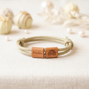 Könnte beinhalten: Ein hellgrünes Seilarmband mit zwei rechteckigen Holzperlen. Eine Perle ist mit dem Namen "Emilia" graviert, die andere zeigt ein Regenbogen-Design. Das Armband ist an den Enden geknotet.