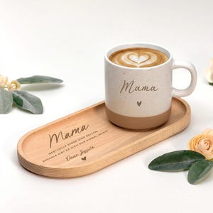 Op de afbeelding: Een witte keramische mok met het woord "Mama" en een hart, gevuld met koffie en latte art, staat op een houten dienblad. Op het dienblad staat het woord "Mama" en andere tekst gegraveerd. De mok en het dienblad staan op een wit oppervlak.