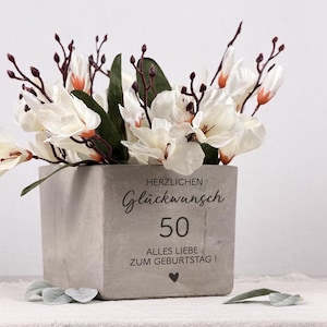 May include: A square, gray concrete planter filled with white and orange flowers and dark brown stems. The planter has the text "HERZLICHEN Glückwunsch 50 ALLES LIEBE ZUM GEBURTSTAG!" and a heart symbol.