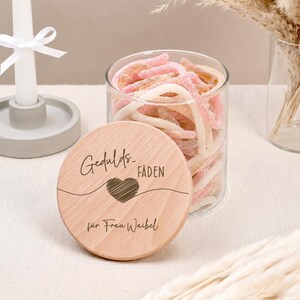 Könnte beinhalten: Ein klares Glasgefäß gefüllt mit rosa und weißen Weingummischnüren. Der runde Holzdeckel ist mit dem Text Gedulds-FÄDEN, einem Herzsymbol und dem Text für Frau Waibel graviert und steht neben einer weißen Kerze.