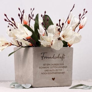May include: A square, light gray concrete planter filled with white and orange-tipped flowers and green leaves. The planter has the word "Freundschaft" and a quote in German. A small heart is below the quote. The arrangement is on a white surface.