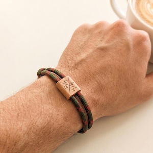 Könnte beinhalten: Ein Armband mit einer doppelt geführten, olivgrün und rot gemusterten Kordel und einer rechteckigen Holzperle. Die Perle ist mit einem Kreuzmuster, dem Buchstaben O, der Zahl 28, dem Buchstaben N und einem kleinen Ankersymbol graviert.