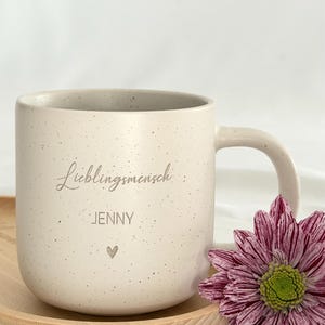 May include: A speckled off-white ceramic mug with a curved handle. The mug has the word "Lieblingsmensch" and the name "Jenny" printed on it, along with a small heart symbol. A purple and white flower rests beside the mug.