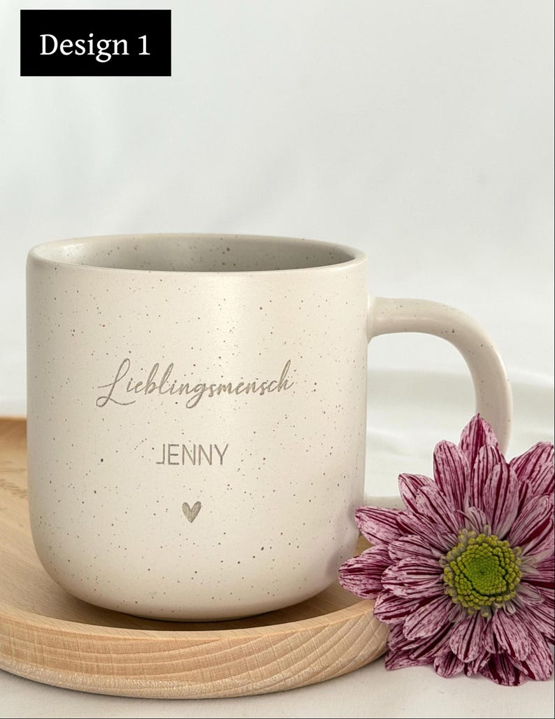 May include: A speckled off-white ceramic mug with a curved handle. The mug has the word "Lieblingsmensch" and the name "Jenny" printed on it, along with a small heart symbol. A purple and white flower rests beside the mug.