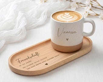 Pode incluir: Uma caneca de cerâmica branca e bege salpicada com o nome Vanessa e um pequeno coração impresso na lateral, sobre uma bandeja oval de madeira gravada com o texto alemão Freundschaft ist ein Zauber, der schwere Zeiten leichter und gute Zeiten noch schöner macht.