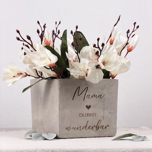 Op de afbeelding: Een vierkante, grijze betonnen plantenbak gevuld met witte bloemen en groene bladeren. De plantenbak heeft de woorden "Mama, Du bist wunderbar" gegraveerd op de voorkant. De bloemen hebben een vleugje oranje aan de basis van de bloemblaadjes.