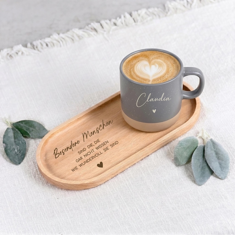 Puede incluir: Una taza de caf&eacute; gris moteada con el nombre "Claudia" y un dise&ntilde;o de coraz&oacute;n, llena de un caf&eacute; con leche con un dise&ntilde;o en forma de coraz&oacute;n, descansa sobre una bandeja de madera. La bandeja tiene texto grabado y un s&iacute;mbolo de coraz&oacute;n.