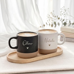 Könnte beinhalten: Zwei Keramiktassen mit Henkel, eine in Schwarz und eine in Weiß, beide mit gesprenkelter Textur und einem beigen Boden. Die schwarze Tasse ist mit dem Namen Oliver und einem kleinen Herz bedruckt, die weiße Tasse mit dem Namen Natalie und Herz.