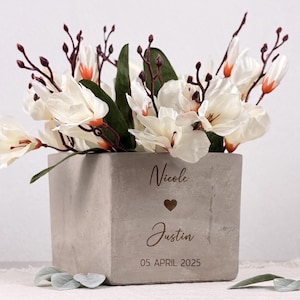 Könnte beinhalten: Ein quadratischer, grauer Betonblumentopf gefüllt mit weißen und orangefarbenen Blumen und grünen Blättern. Der Topf ist mit den Namen "Nicole" und "Justin", einem Herzsymbol und dem Datum "05. APRIL 2025" graviert.