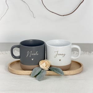 Puede incluir: Dos tazas de cer&aacute;mica moteada, una gris y otra blanca, cada una con un nombre y un detalle de coraz&oacute;n. Las tazas est&aacute;n sobre una bandeja de madera con una flor y hojas. La taza gris dice "Nicole" y la blanca "Jenny".