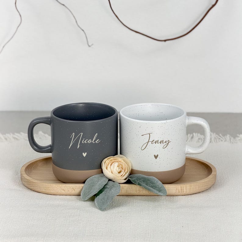 Puede incluir: Dos tazas de cer&aacute;mica moteada, una gris y otra blanca, cada una con un nombre y un detalle de coraz&oacute;n. Las tazas est&aacute;n sobre una bandeja de madera con una flor y hojas. La taza gris dice "Nicole" y la blanca "Jenny".