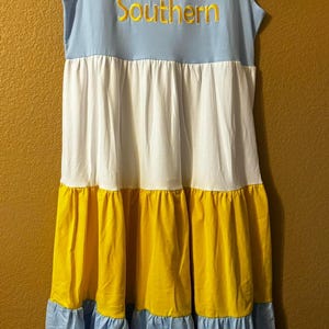 May include: A light blue, white, and yellow tiered dress with short ruffle sleeves. The dress has the word "Southern" embroidered on the front in gold thread.