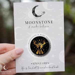 May include: A gold-coloured phone grip with a black background and a silhouette of a stag with antlers and a crown. The text "Moonstone & Make Believe" is written around the edge of the grip. The text "Kindle Grip" and "Life is too short to read a bad book." is written below the grip.