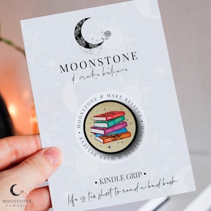 May include: A round, white sticker with a black border and a stack of colourful books. The sticker says "Moonstone & Make Believe" around the edge. The text "Kindle Grip" and "Life is too short to read a bad book." are printed below the sticker.