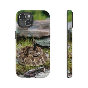 May include: A phone case featuring a coiled rattlesnake in its natural habitat. The snake is brown and tan with a black tail, resting on moss-covered rocks. The case has a black border and a camera cutout.