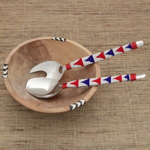 Puede incluir: Un cuenco de ensalada de madera con detalles decorativos en blanco y negro, junto con un juego de servir. Los utensilios para servir tienen cabezas plateadas y mangos adornados con patrones de cuentas rojas, blancas y azules. El conjunto descansa sobre una superficie texturizada de tono neutro.