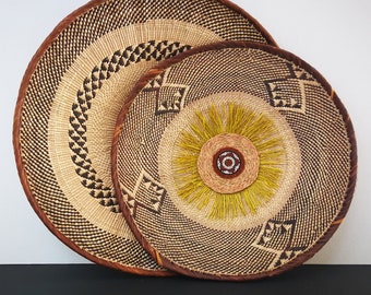 May include: Two woven straw baskets with a brown and black geometric pattern. The smaller basket is nested inside the larger basket and has a yellow flower design in the center.