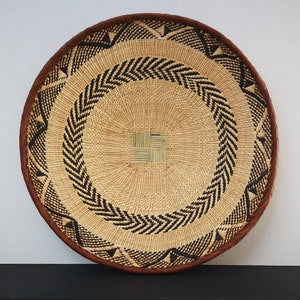 May include: Round woven basket with a natural beige center, accented with black chevron and triangle patterns. The rim is a dark brown, creating a contrasting border. The basket is likely used for decorative or storage purposes.