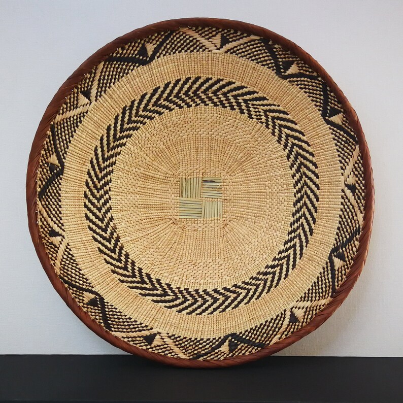 May include: Round woven basket with a natural beige center, accented with black chevron and triangle patterns. The rim is a dark brown, creating a contrasting border. The basket is likely used for decorative or storage purposes.