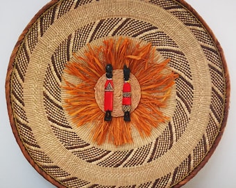 May include: A round woven wall hanging with a brown and beige pattern. The center of the hanging features a cluster of orange fibers with two small wooden figures wearing red and black clothing.