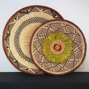May include: Two round woven wall baskets. The larger basket has a light brown and black geometric pattern with a dark brown rim. The smaller basket has a similar pattern with a green and brown decorative center. Home decor.