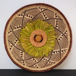 May include: Round woven wall decor featuring a central floral design. The outer ring is a dark brown, with a lighter woven pattern inside. The center has a circular design with green, brown, and red accents. The overall design is natural and earthy.