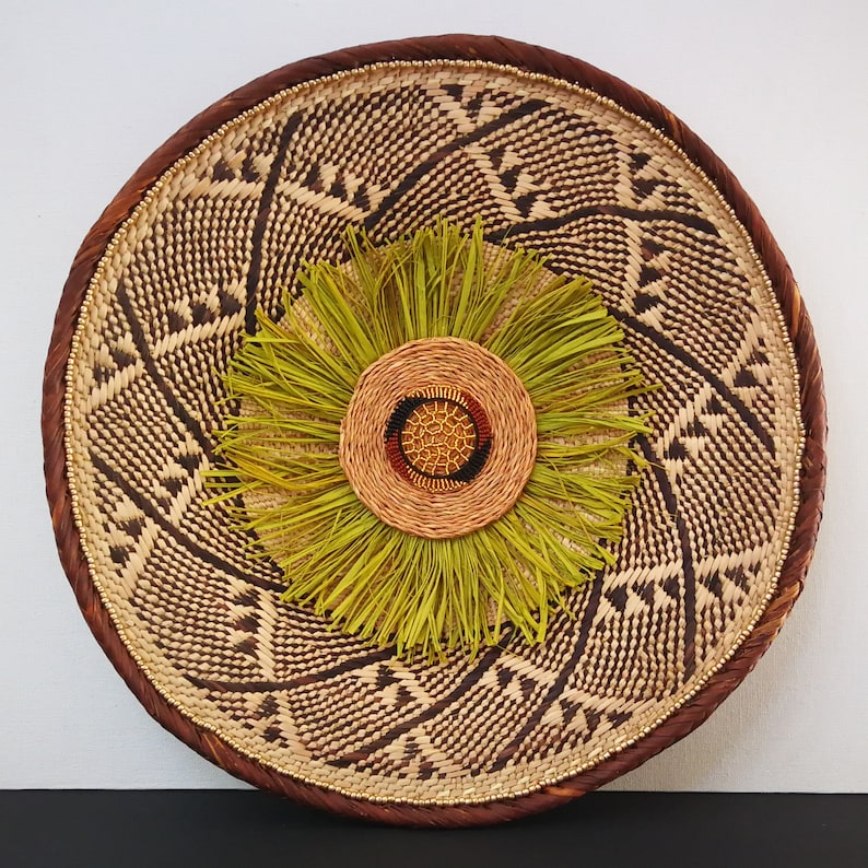May include: Round woven wall decor featuring a central floral design. The outer ring is a dark brown, with a lighter woven pattern inside. The center has a circular design with green, brown, and red accents. The overall design is natural and earthy.