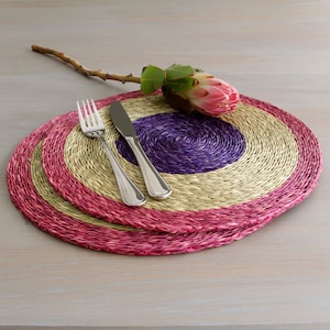Puede incluir: Dos manteles individuales redondos tejidos con rayas rosas, beige y moradas. Un tenedor y un cuchillo están colocados encima de los manteles individuales.