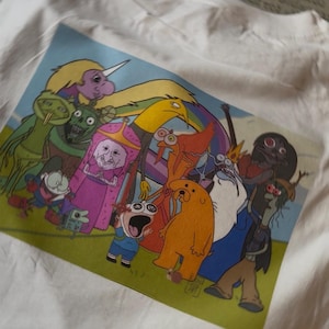Puede incluir: Camiseta blanca con una colorida impresión gráfica de varios personajes de dibujos animados. El diseño presenta figuras diversas en una composición colorida, sobre un fondo azul y verde. La camiseta es de tejido blanco suave.