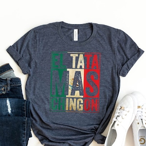May include: A dark blue t-shirt with a graphic print of the text "El Tata Mas Chingon" in a colorful block style reminiscent of the Mexican flag. The shirt is shown on a white background with a pair of blue jeans and white sneakers.