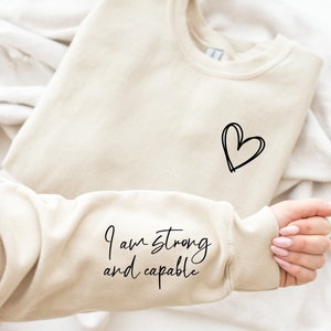 May include: A beige sweatshirt with the text "I am strong and capable" on the sleeve and a simple heart design on the front.
