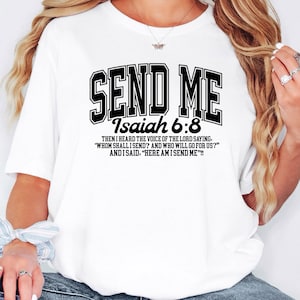 May include: White t-shirt with the text "SEND ME" in a black, collegiate-style font. Below the text is "Isaiah 6:8" and a quote from the Bible. The shirt is a casual, everyday wear item.