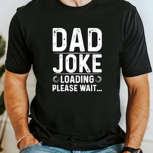 May include: Black t-shirt with the text "DAD JOKE" in white, distressed font. Below the text is "LOADING PLEASE WAIT..." in a smaller, stylized font. The shirt is short-sleeved and appears to be made of a soft material.