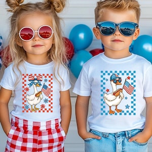 May include: Two children wearing white t-shirts with goose graphics. The goose illustrations hold American flags and are set against a red, white, and blue checkered background. One child wears red sunglasses and the other wears blue sunglasses.