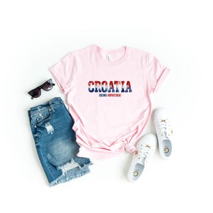 K&ouml;nnte beinhalten: Ein hellrosa T-Shirt mit dem Wort "CROATIA" in Rot, Wei&szlig; und Blau und dem Text "IDEMO HRVATSKA!". Das Shirt wird mit Jeansshorts, Sonnenbrille und wei&szlig;en Turnschuhen kombiniert.
