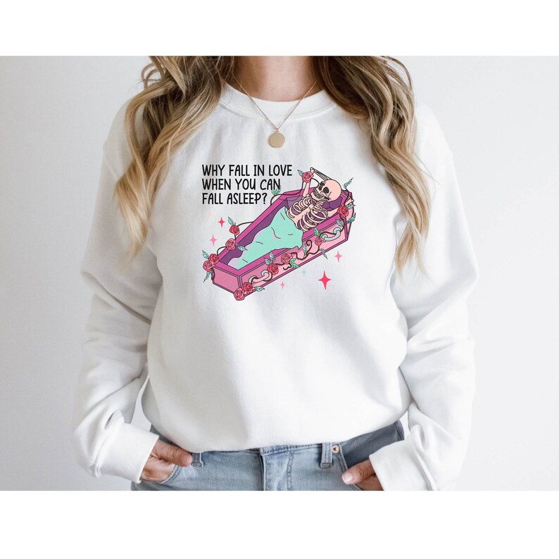 May include: White sweatshirt with a pink and green coffin with a skeleton inside. The text on the sweatshirt reads "Why fall in love when you can fall asleep?"