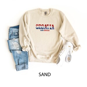 May include: A sand-colored sweatshirt with the word "CROATIA" in red, white, and blue, with the text "IDEMO HRVATSKA!" below it. The sweatshirt is paired with blue jeans and white sneakers.