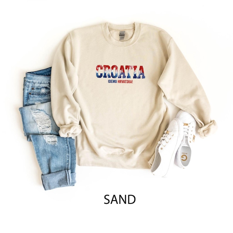 May include: A sand-colored sweatshirt with the word "CROATIA" in red, white, and blue, with the text "IDEMO HRVATSKA!" below it. The sweatshirt is paired with blue jeans and white sneakers.