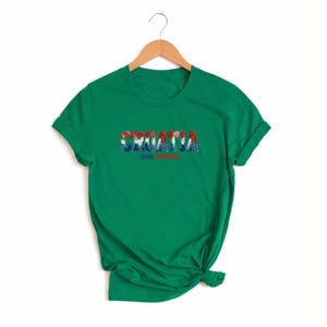 K&ouml;nnte beinhalten: Ein gr&uuml;nes T-Shirt mit dem Wort "CROATIA" in einem farbenfrohen, von der Flagge inspirierten Design auf der Brust. Darunter steht "IGEMU ZAGREB". Das T-Shirt h&auml;ngt an einem Holzb&uuml;gel.