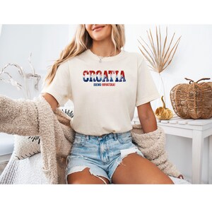 K&ouml;nnte beinhalten: Cremefarbenes T-Shirt mit dem Wort "CROATIA" in Rot, Wei&szlig; und Blau, darunter "IDEMO HRVATSKA!". Das Shirt wird mit Jeansshorts getragen. Das Gesamtthema ist Reisen und Nationalstolz.