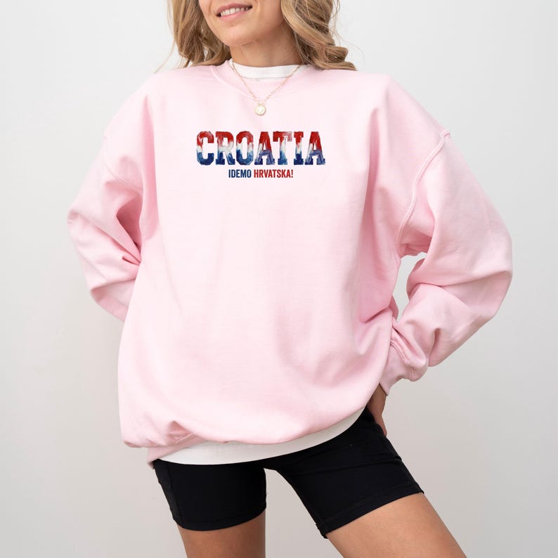 May include: A light pink sweatshirt with the word "CROATIA" in red, white, and blue lettering across the chest. Below the word "CROATIA" is the text "IDEMO HRVATSKA!" in white. The sweatshirt is paired with black shorts.