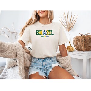 以下が含まれることがあります： クリーム色のTシャツ。緑と黄色で「BRAZIL」の文字、青で「VAMOS BRASIL！」のフレーズがプリントされています。デニムショートパンツと合わせています。全体的にカジュアルでリラックスした雰囲気です。