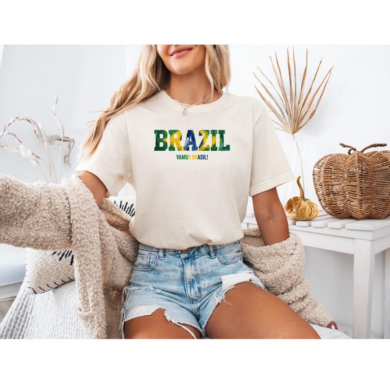 以下が含まれることがあります： クリーム色のTシャツ。緑と黄色で「BRAZIL」の文字、青で「VAMOS BRASIL！」のフレーズがプリントされています。デニムショートパンツと合わせています。全体的にカジュアルでリラックスした雰囲気です。