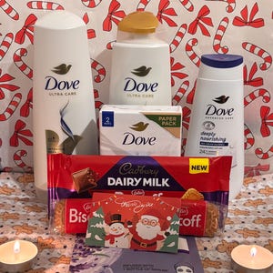 Puede incluir: Una colección de productos de baño Dove, chocolate Cadbury Dairy Milk, galletas Lotus Biscoff y una mascarilla facial. Los artículos están dispuestos sobre un fondo festivo con bastones de caramelo y lazos, con velas encendidas.