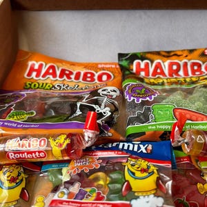 May include: A box filled with various bags of Haribo candies. The bags feature Halloween-themed graphics, including skeletons and monsters. Other candies include Haribo Roulette and Fruit Juice gummies. The packaging is colorful, with orange, green, and blue hues.